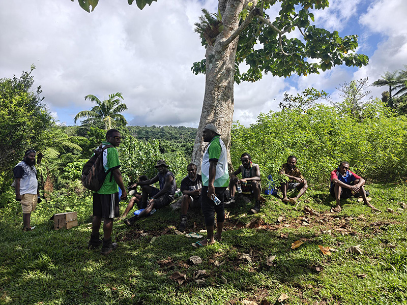 Ni-Vanuatu CCA rangers standing and sitting by a tree on a hilltop surrounded by tropical forest. 